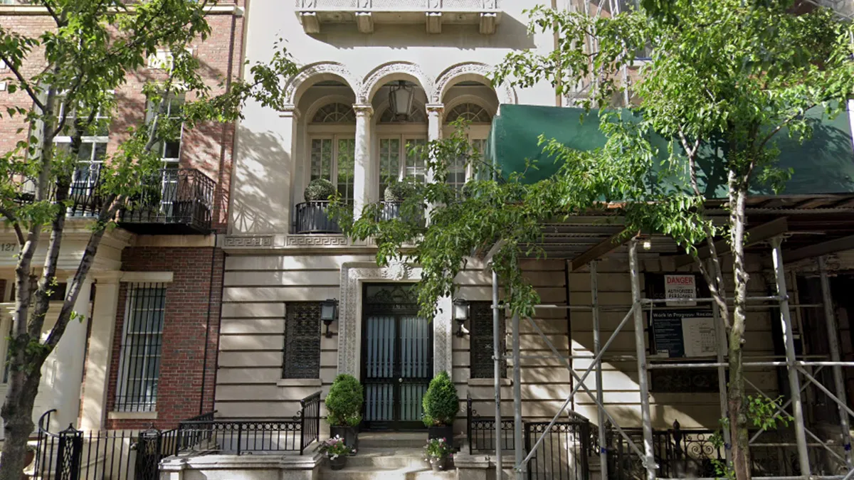 Three-story townhouse with decorative arches on the second floor, a black entry door, and potted plants, with scaffolding and a green awning on the right amid trees.