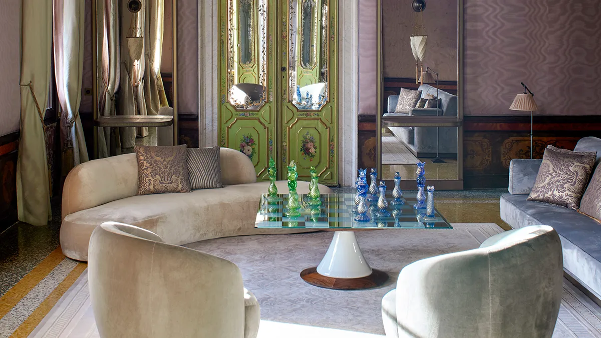 Luxurious living room with a curved beige sofa, decorative cushions, and a glass chessboard centerpiece in front of ornate green doors.