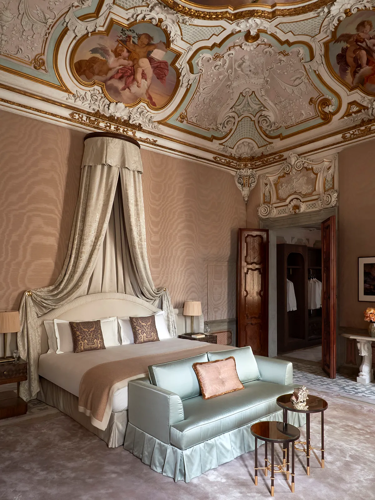 Luxurious bedroom with a canopy bed and draped linen, ornate painted ceiling, and a pale blue loveseat at the foot of the bed.