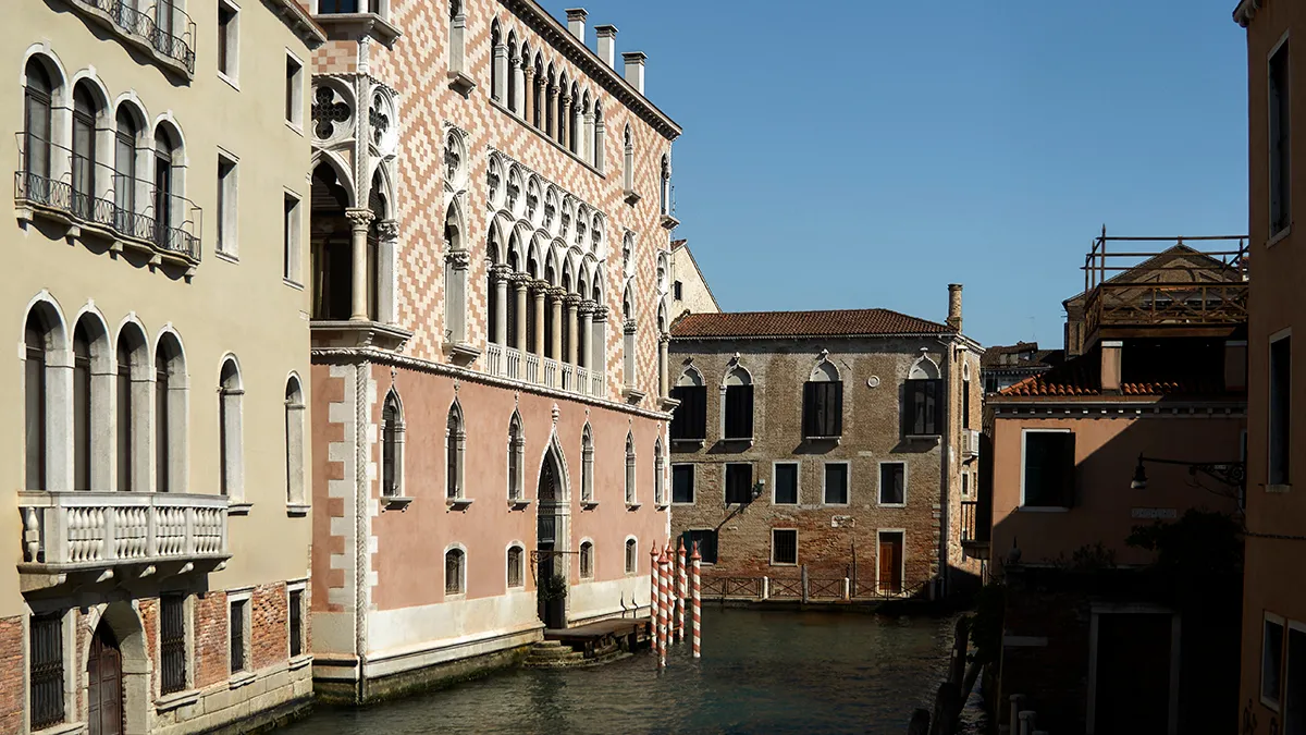 Colorful Venetian buildings line a narrow canal, with striped mooring poles by the water and bright blue sky above.