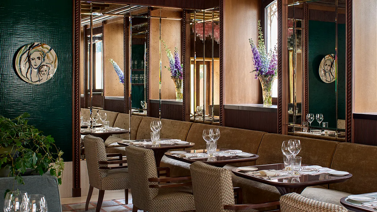Restaurant dining area with round tables set for service, beige chairs, wine glasses, and purple flower arrangements along mirrored walls.