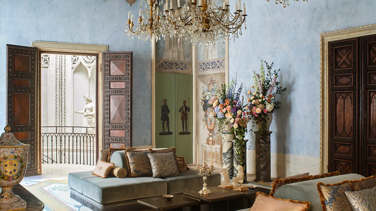 Opulent living room with blue walls, a crystal chandelier, plush velvet couches, and tall floral arrangements in marble vases.