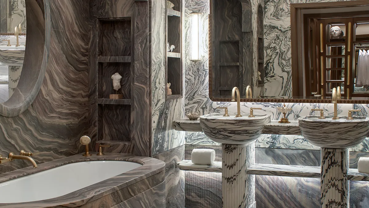 Luxurious marble bathroom with double pedestal sinks, gold faucets, and a veined marble tub.