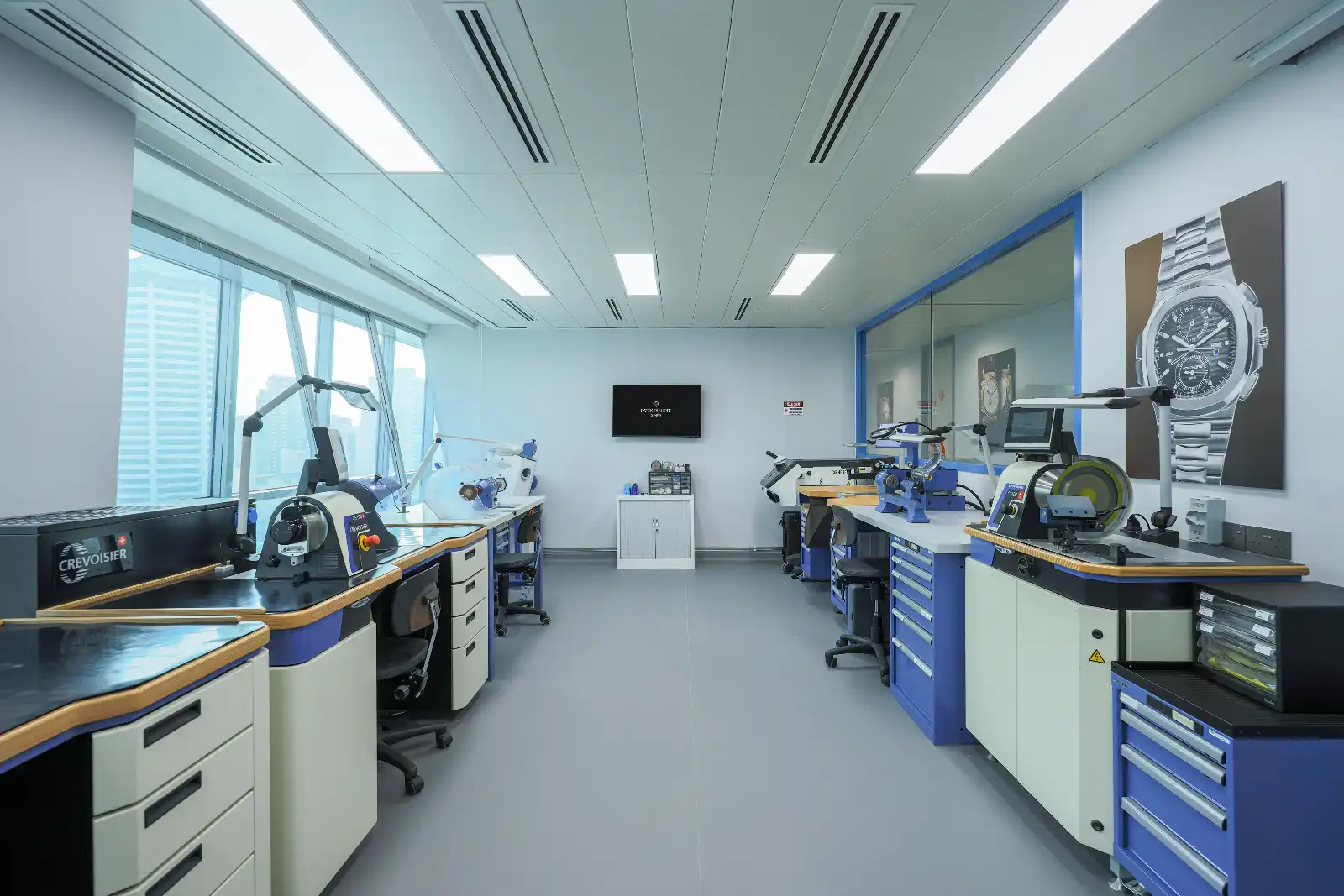 Bright watchmaking workshop with multiple workbenches, precision machines, and blue-and-white cabinets on both sides.