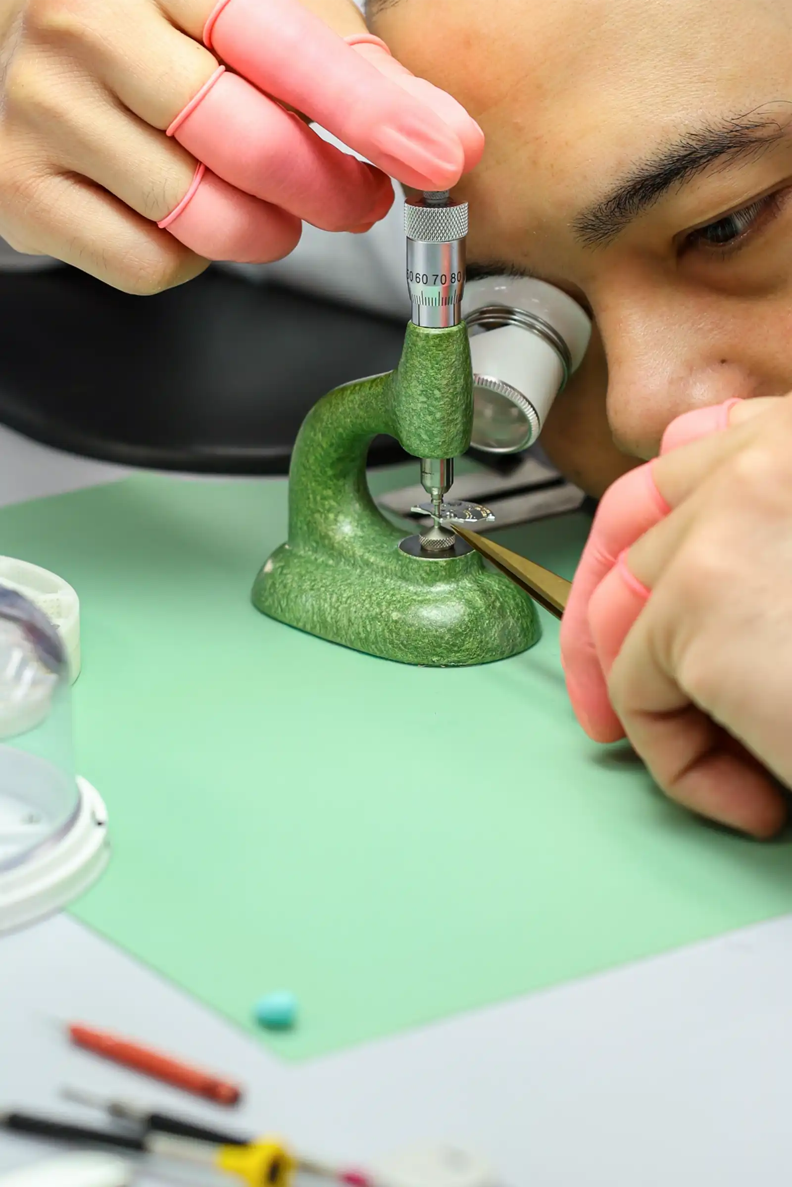 Close-up of gloved hands adjusting a precision micrometer on a small circular metal piece held in a green vise, during detailed work.",