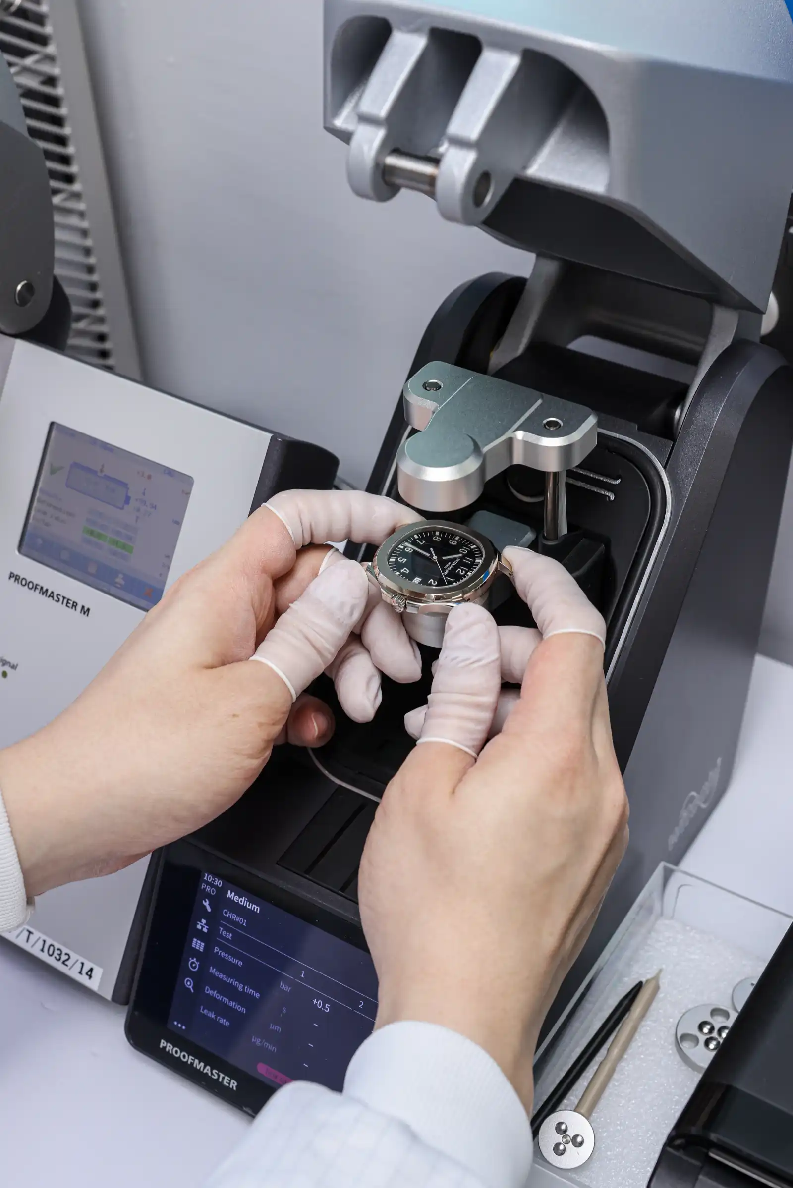 Gloved hands place a wristwatch into a precision testing instrument for quality control.",
