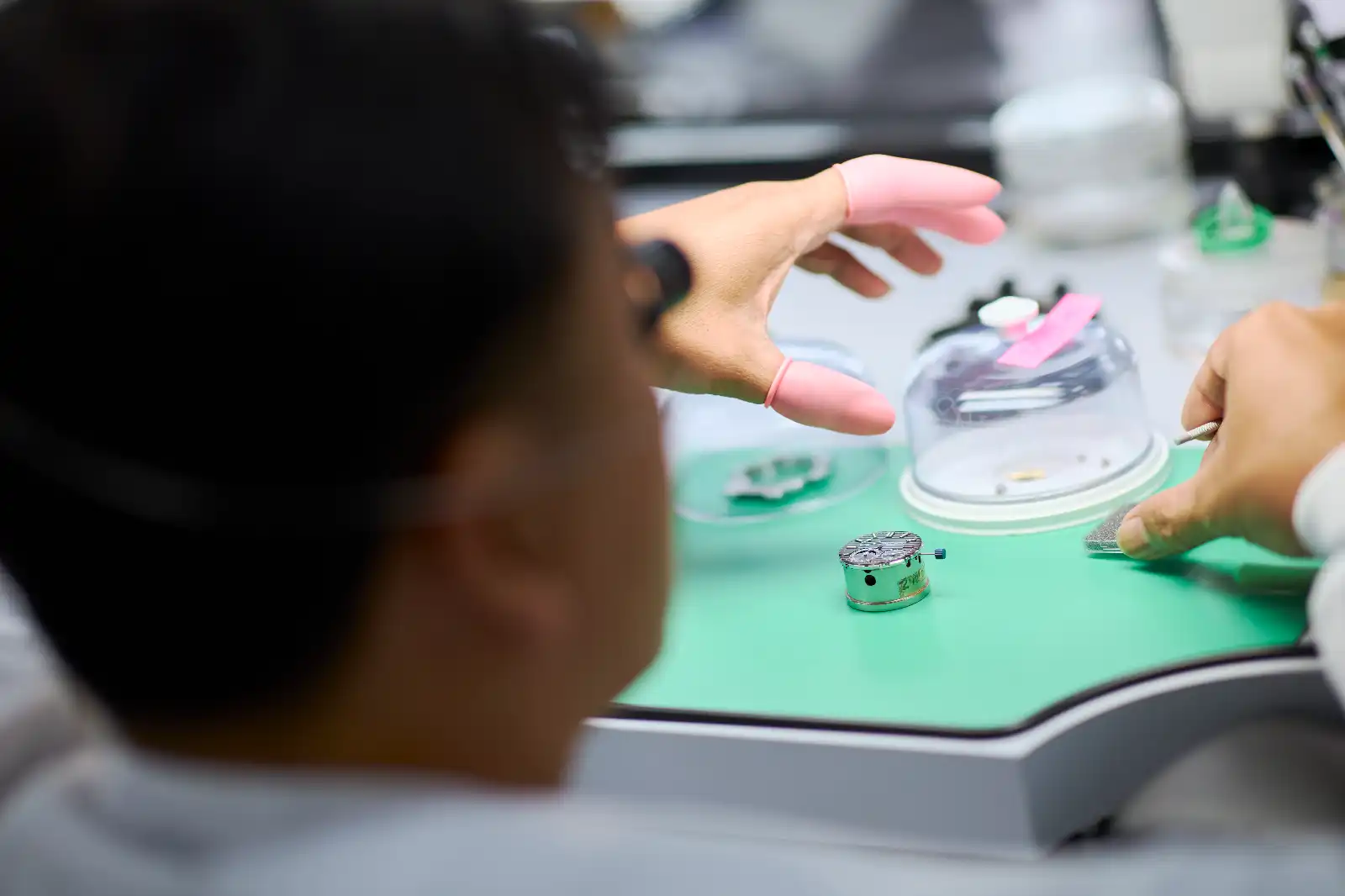 Close-up of a watchmaker's desk with a small watch movement on a green mat and gloved hands handling components under a glass dome nearby