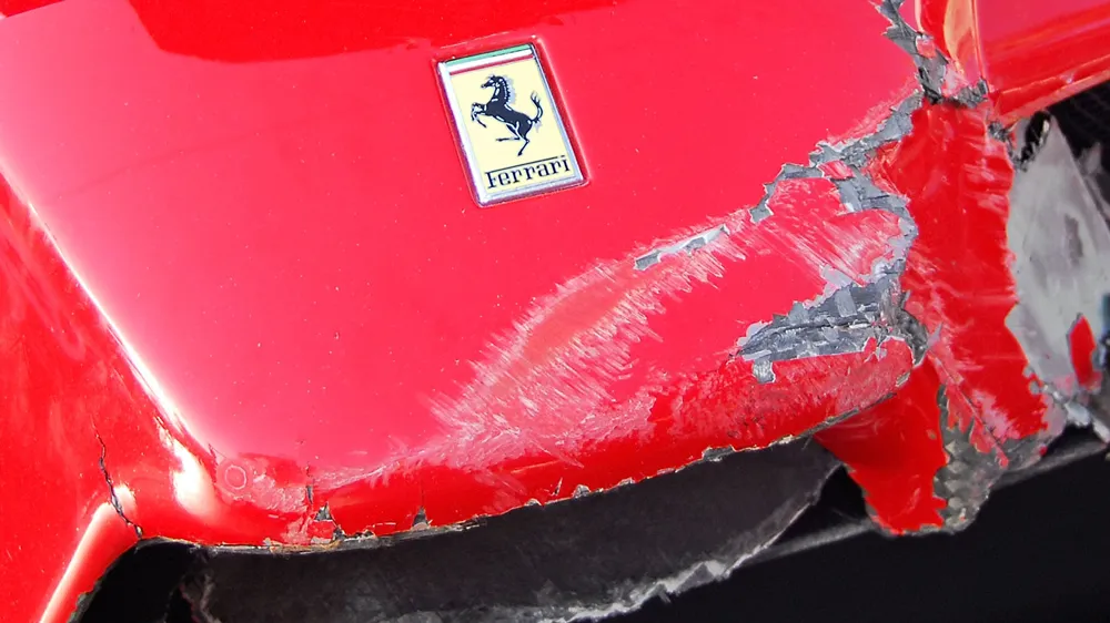 Red Ferrari with significant scraped and ripped bumper damage showing exposed underlying material and peeling paint near the emblem.