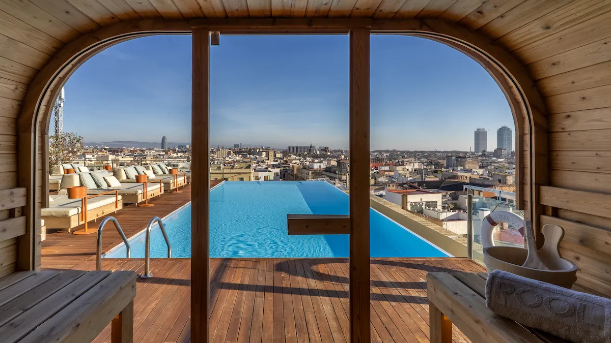 Rooftop pool viewed from a wooden cabana, with lounge chairs and a city skyline in the distance.