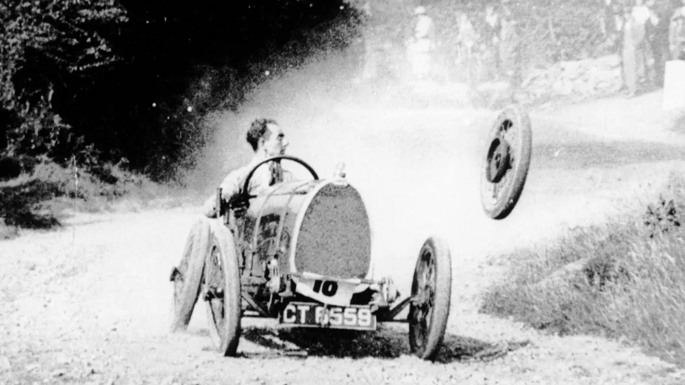 Black-and-white photo of an early 1900s open-cockpit race car on a dusty dirt road, driver steering as a wheel flies off to the right.