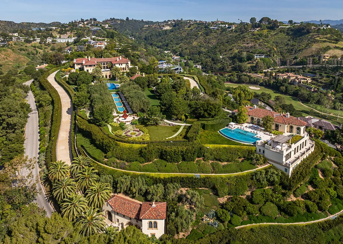 Aerial view of a large Mediterranean-style estate on a hillside with multiple swimming pools, terraced gardens, and winding driveways surrounded by lush trees and hedges.