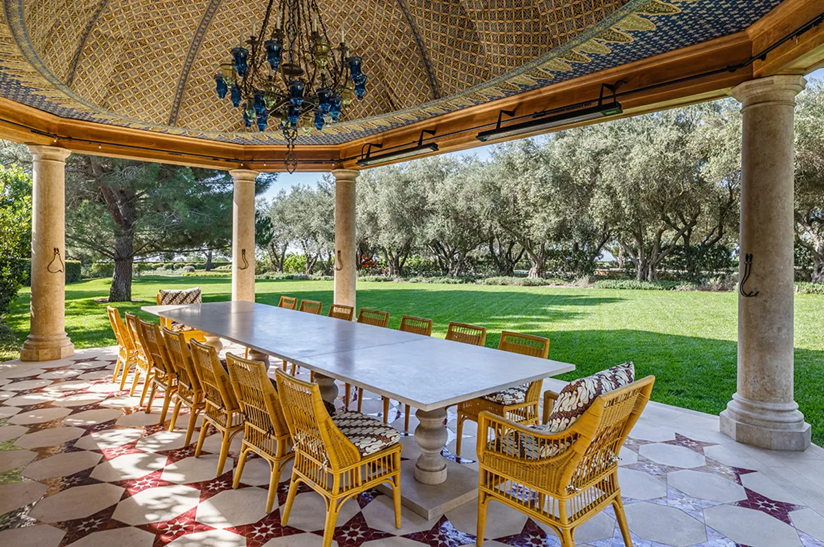 Outdoor covered dining area with a long marble table and wicker chairs, supported by stone columns and a mosaic ceiling.