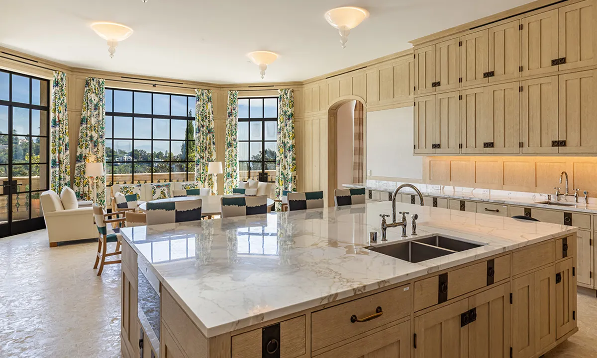 Bright open kitchen with a marble island, light wood cabinets, and a sunny seating area by large windows with floral curtains.