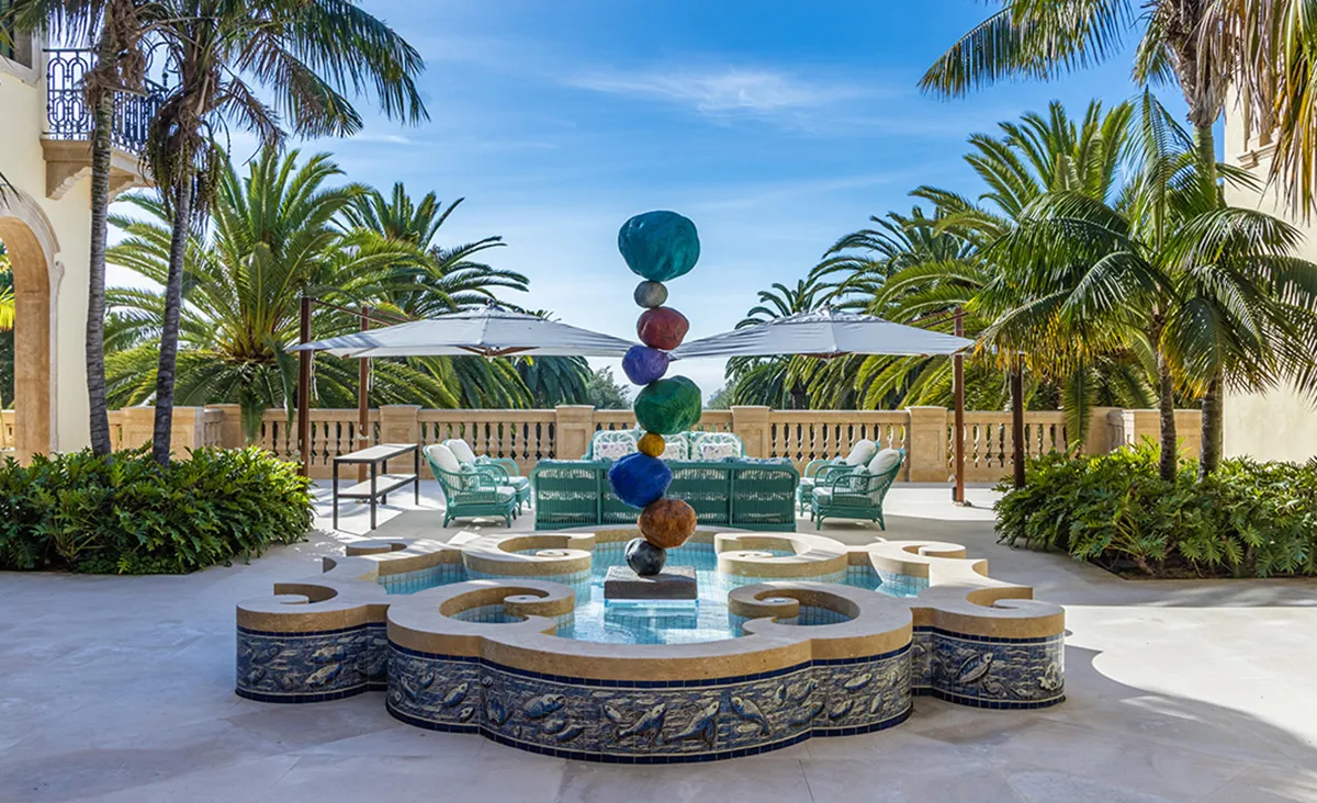 Tiered fountain with stacked colorful spheres at a luxury resort courtyard, surrounded by palm trees and teal seating beneath umbrellas, on a sunny day.