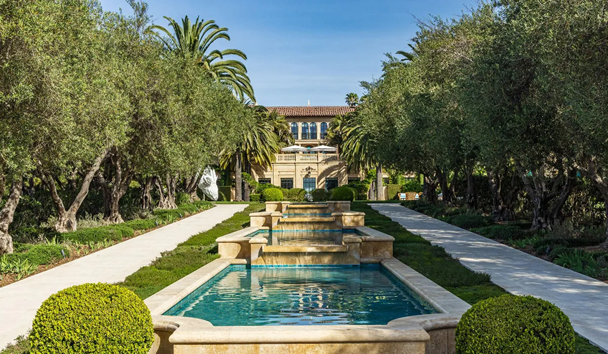 Formal garden with a long cascading water fountain and symmetry, leading to a grand villa framed by olive trees and palm trees.