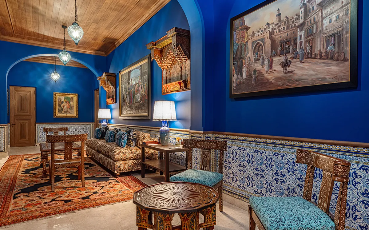Blue-walled living room with Moroccan-inspired furniture, tiled wainscoting, and colorful paintings on the walls.