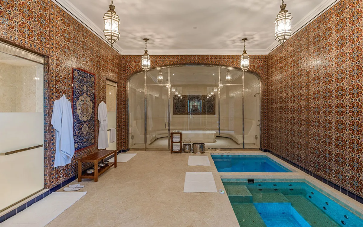 Indoor spa room with ornate tiled walls, robes on a bench, and a glass-enclosed relaxation area in the back.