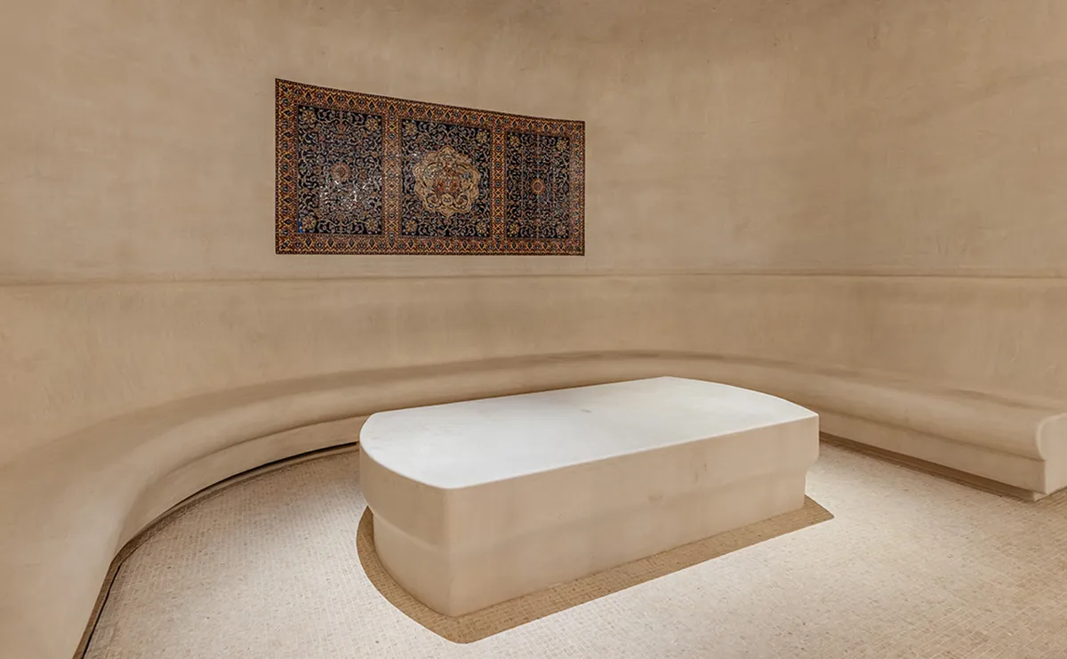 Minimalist beige room with curved plaster walls and a white rounded bench; a patterned rug hangs on the wall above.