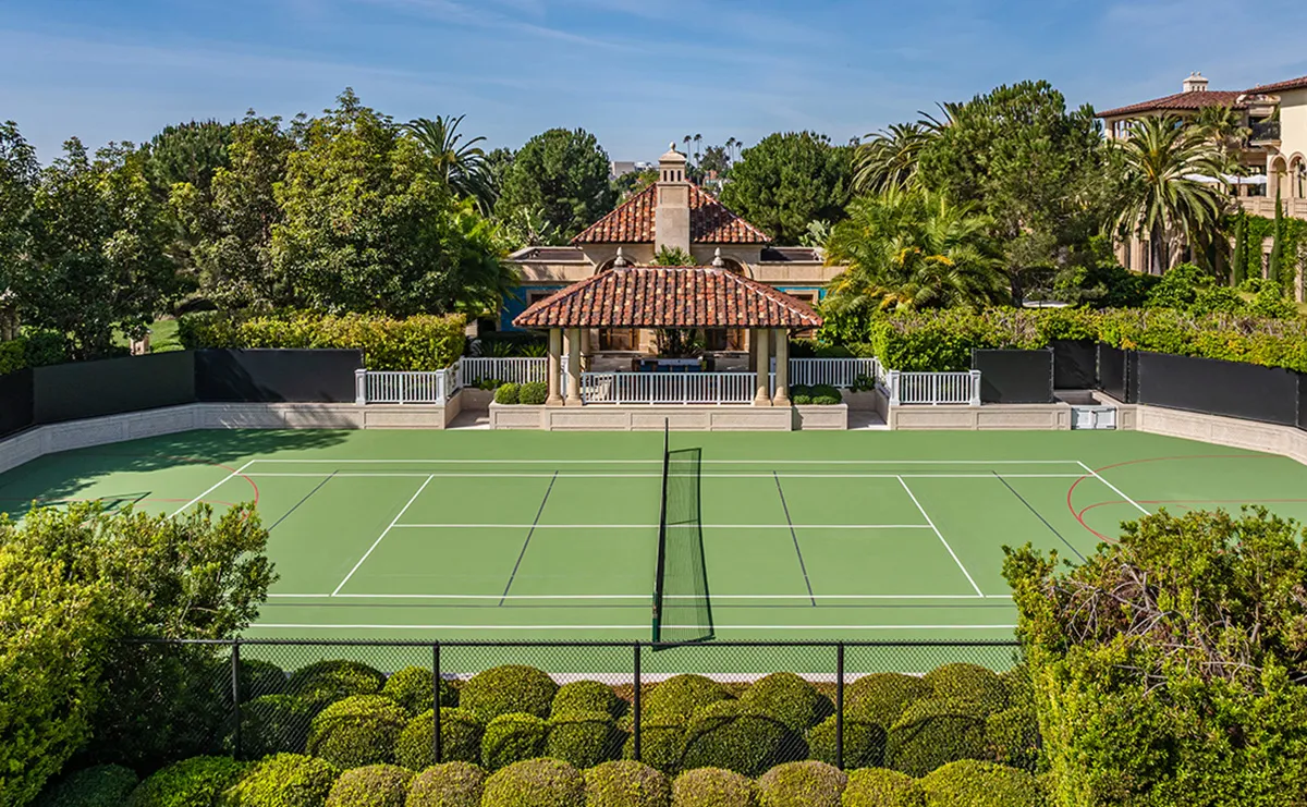 An outdoor tennis court with a central net, surrounded by hedges and a black fence, and a tiled pavilion with a red roof in the background.