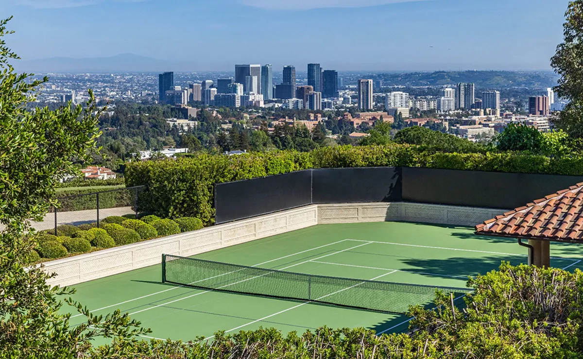 Green tennis court with a net, surrounded by hedges, overlooking a city skyline.