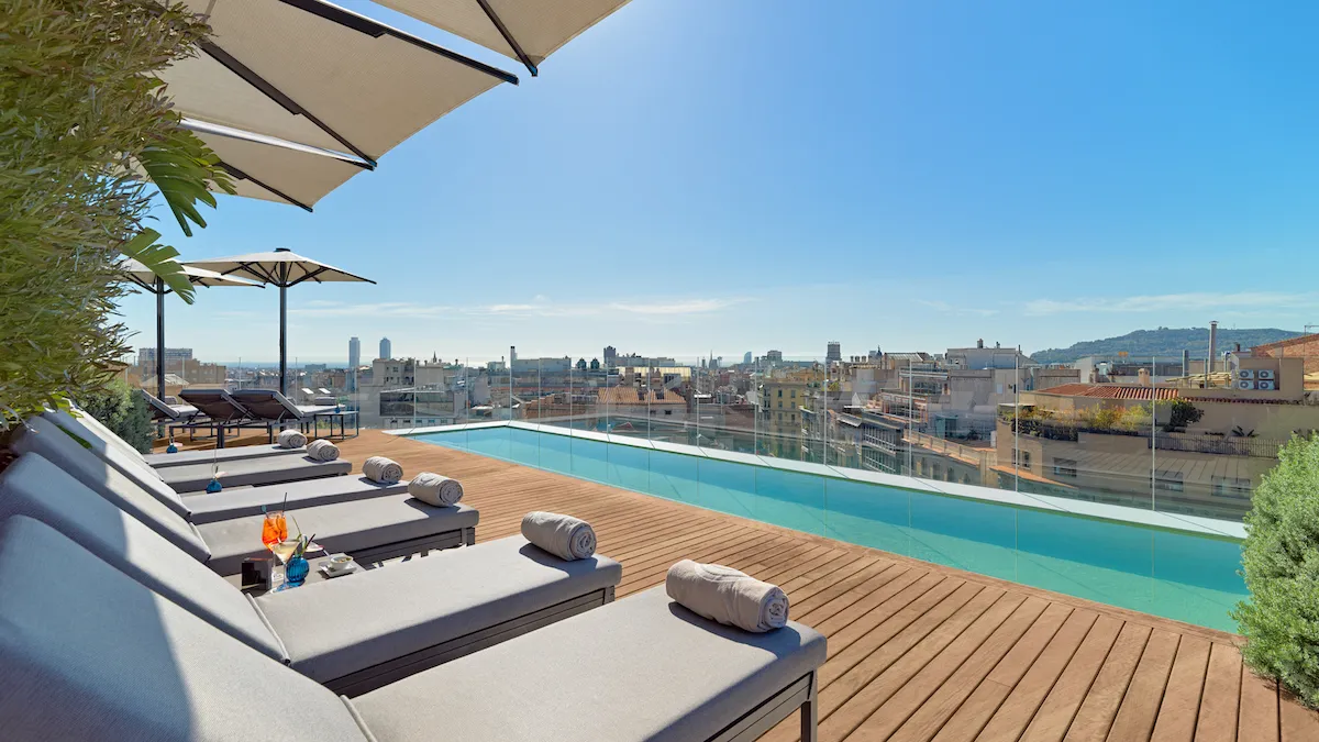 Rooftop pool deck with loungers, umbrellas, and towels, overlooking a city skyline under a clear blue sky.