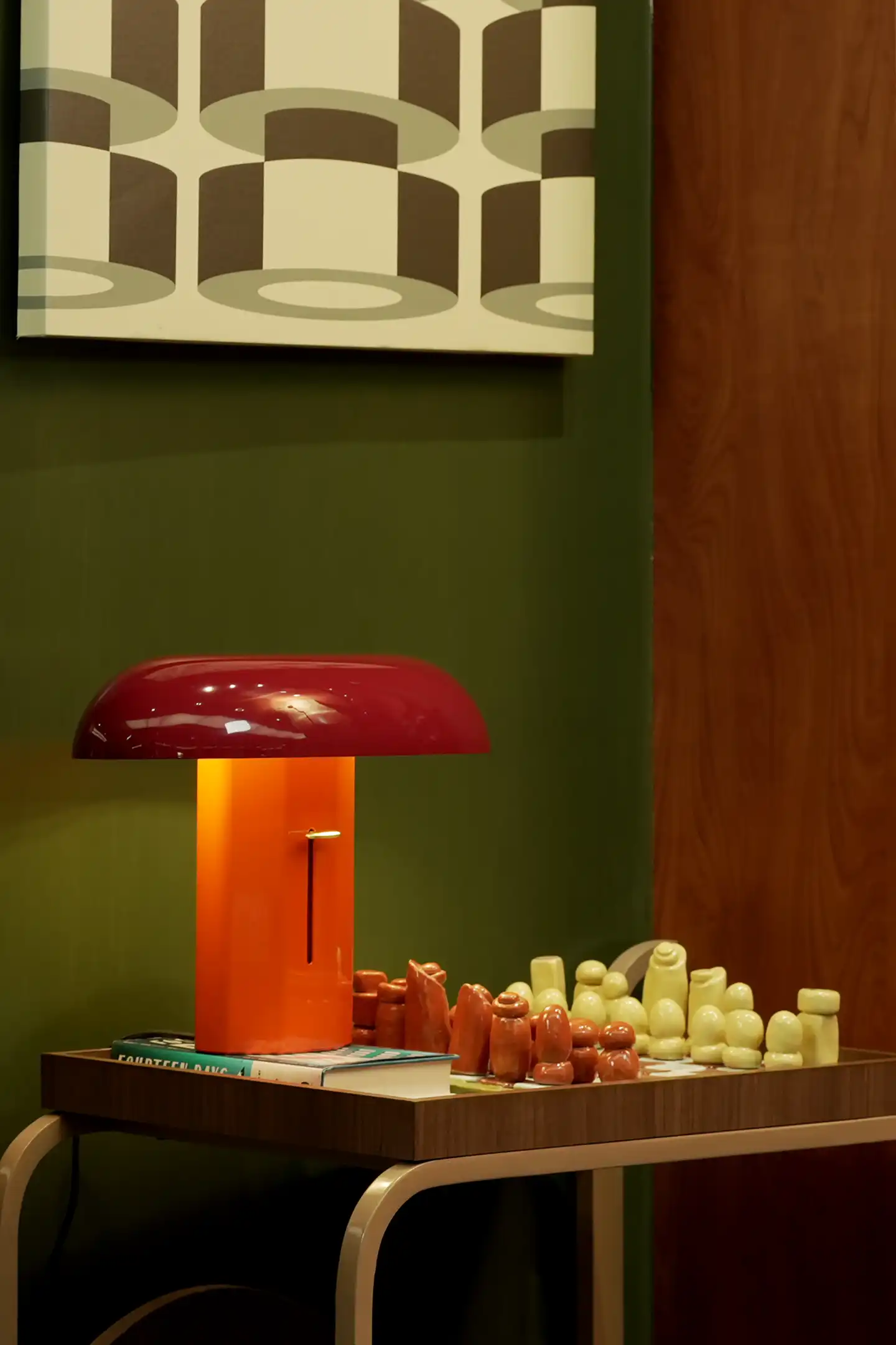 Desk scene with an orange lamp and a red lampshade atop a chessboard filled with white and brown chess pieces against a dark green wall and wooden background.