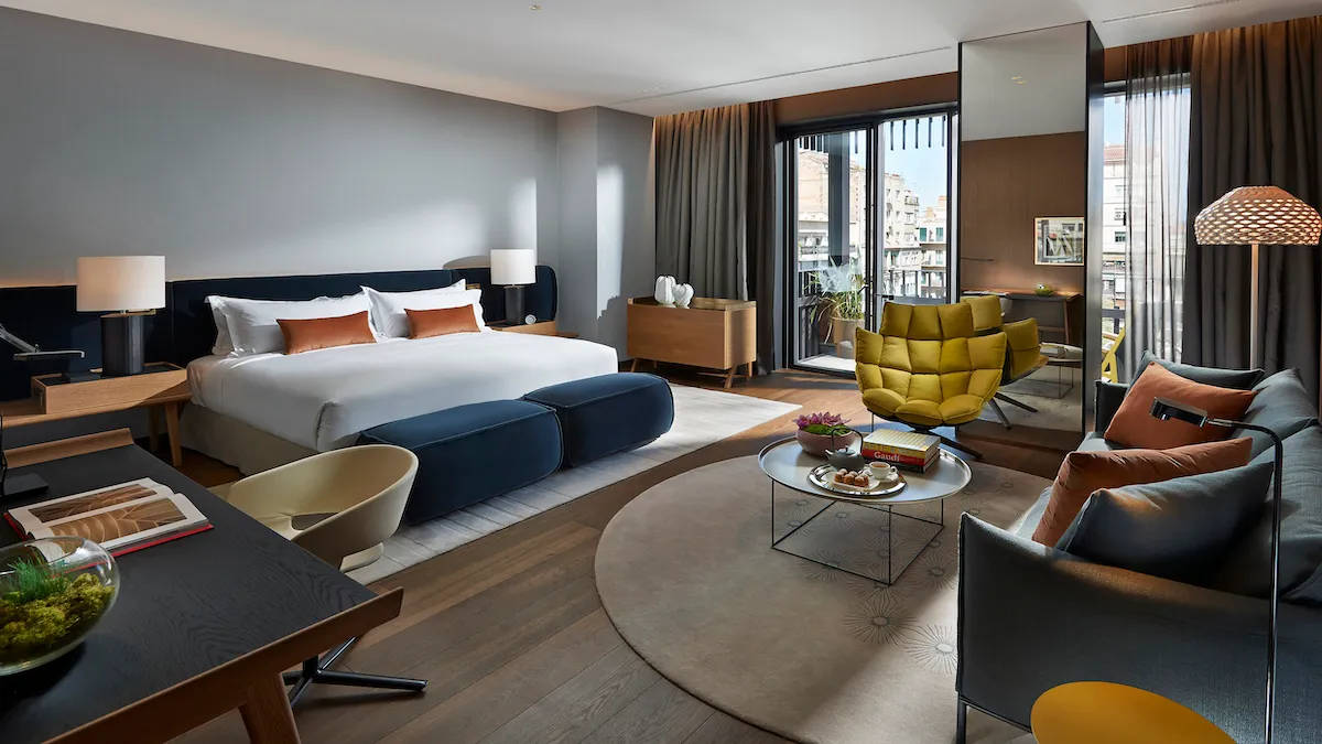 Luxurious hotel suite living area with a king bed, navy headboard, white and orange pillows, and a blue ottoman near the foot of the bed.