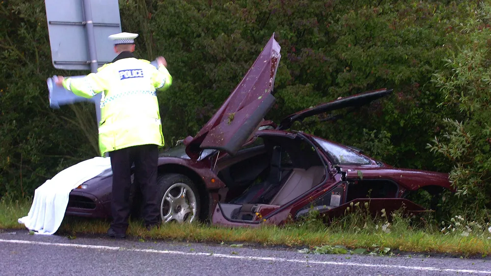 Police officer in a high-visibility jacket stands beside a severely damaged maroon car crashed on the roadside, with a white sheet covering something near the front of the vehicle.