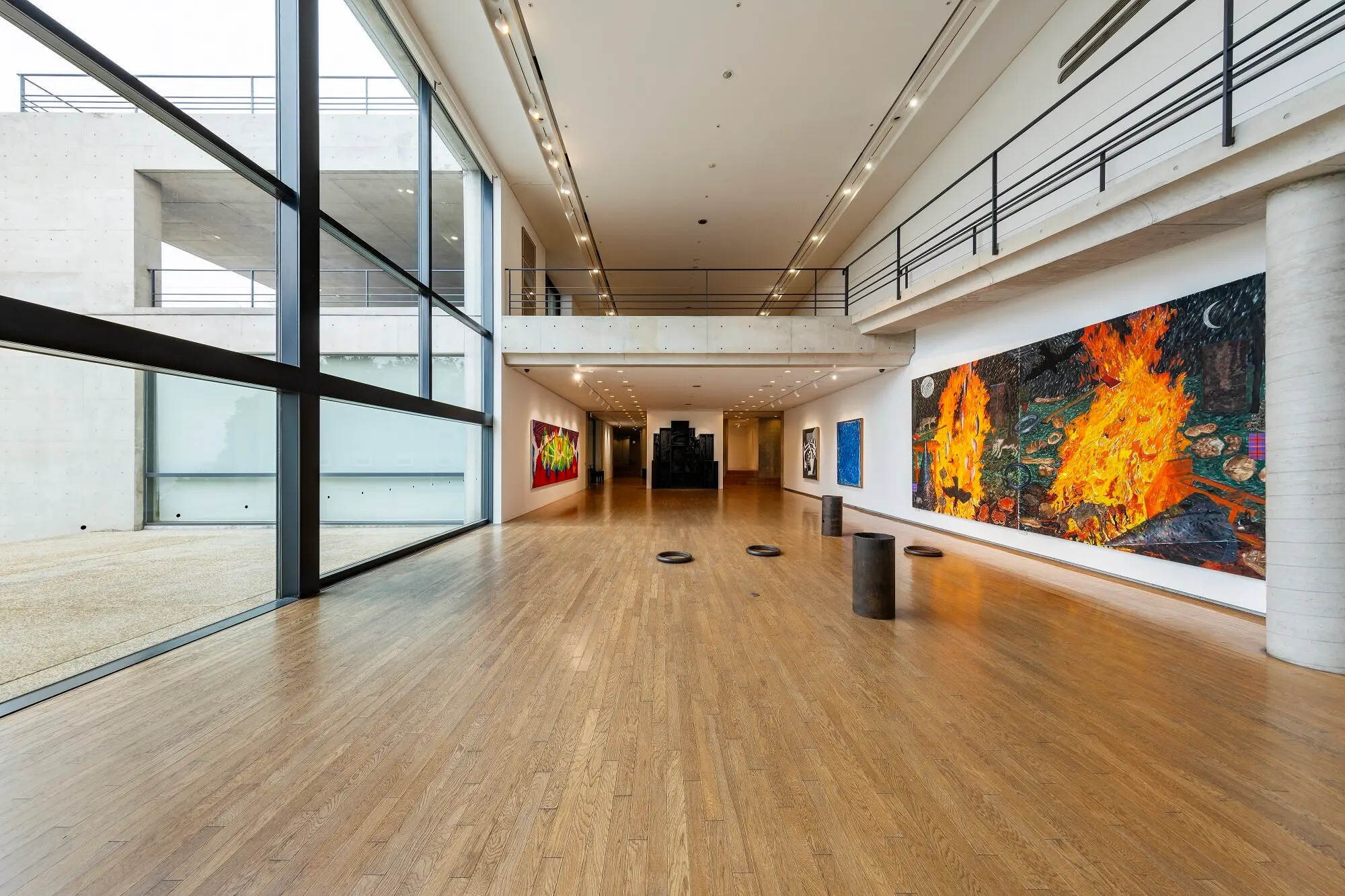 Spacious contemporary art gallery with large glass walls, concrete surfaces, and colorful abstract paintings along the right wall; wooden floor and a mezzanine balcony above.