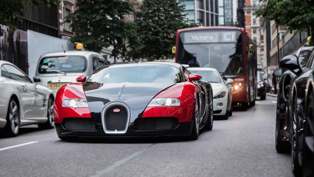 Front view of a red-and-black Bugatti sports car driving in a city street, with a bus and other cars behind.