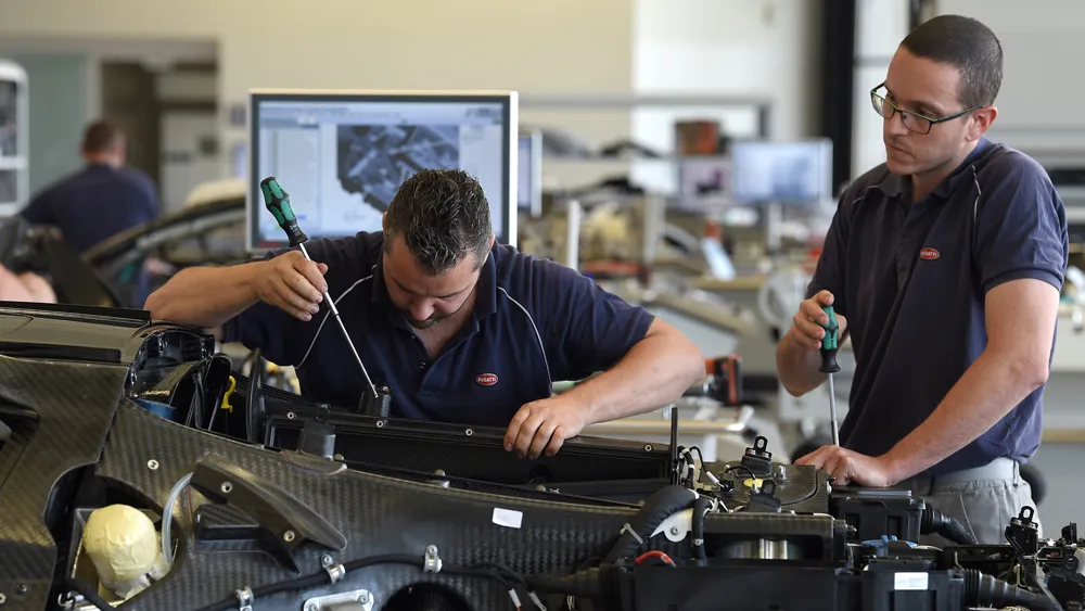 Two technicians in a workshop repair a car engine, one using a screwdriver while the other watches nearby, computer monitor in the background.