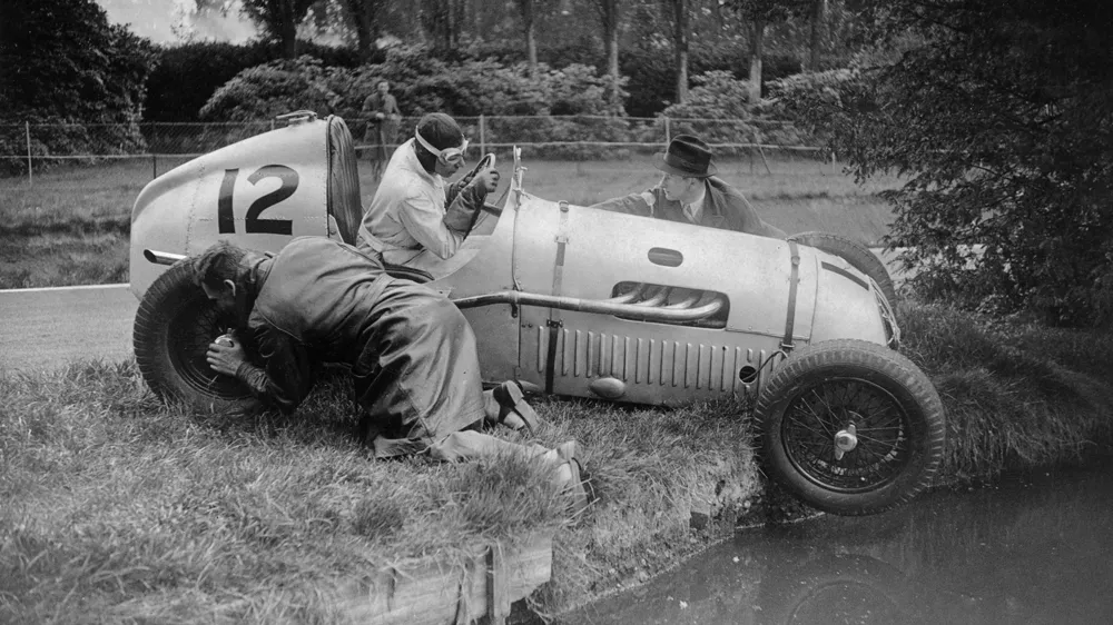 Vintage open-wheel race car with the number 12 overturned on grass beside a canal; three men in period clothing inspect the wrecked car.