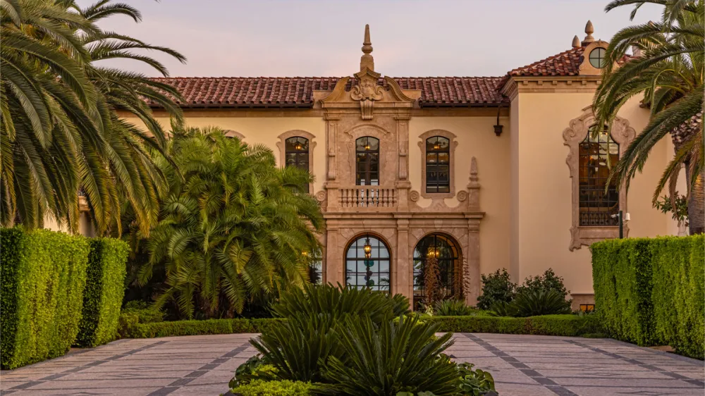 Ornate Mediterranean villa with arched windows, stone facade, and palm-lined courtyard at dusk.