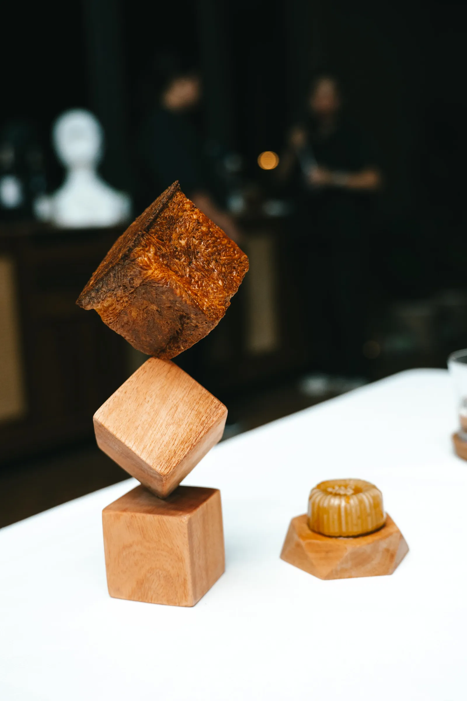 Dark chocolate square perched on a wooden block display, with blurred background and a caramel candy nearby on a white surface.