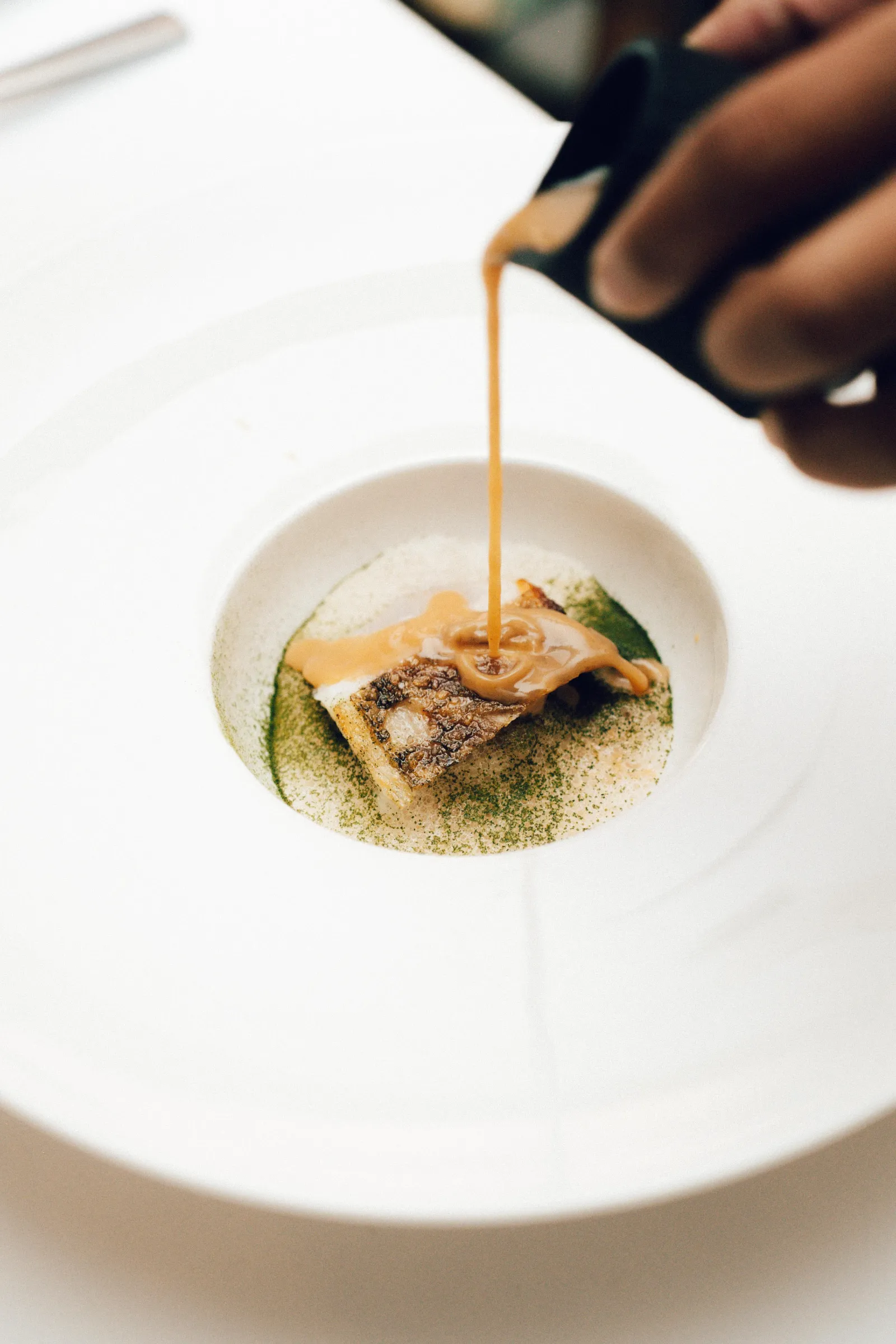 Chef pours a thin orange sauce over a small plated appetizer on a white plate, close-up view.