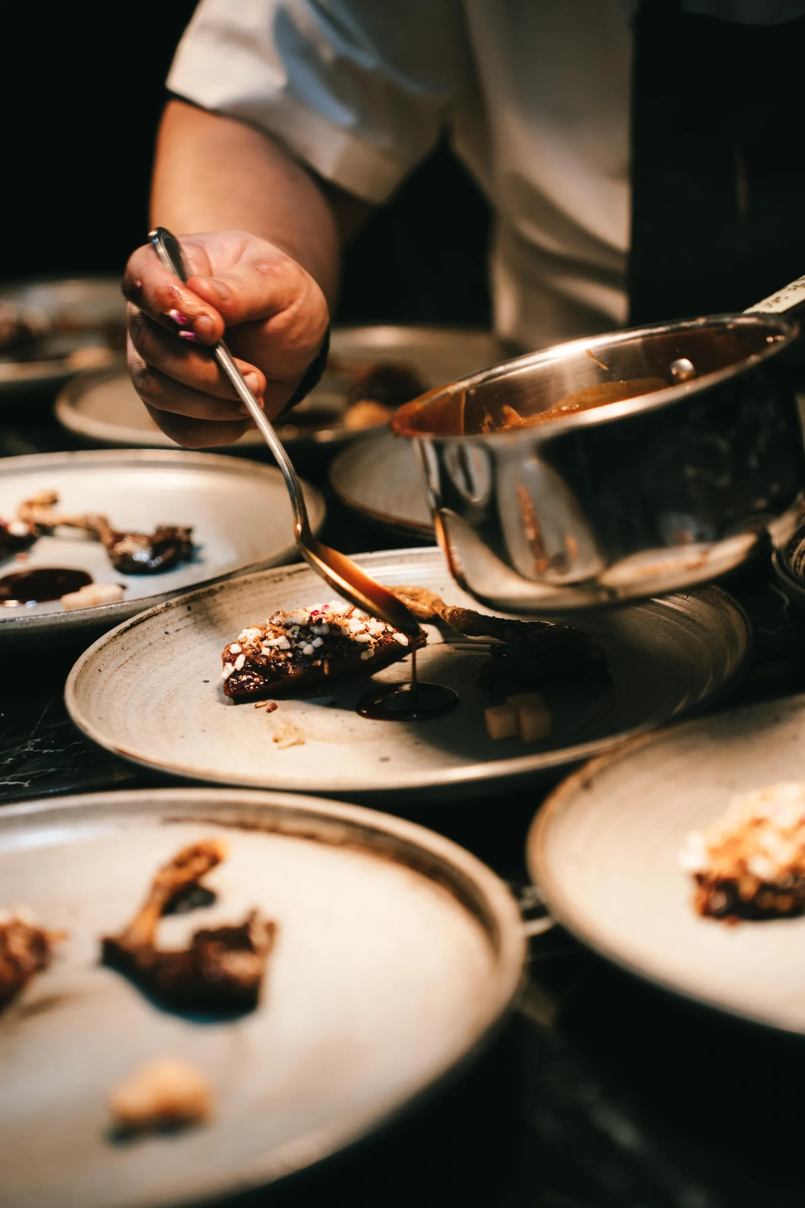 Chef drizzling chocolate sauce onto a plated dessert in a busy kitchen.
