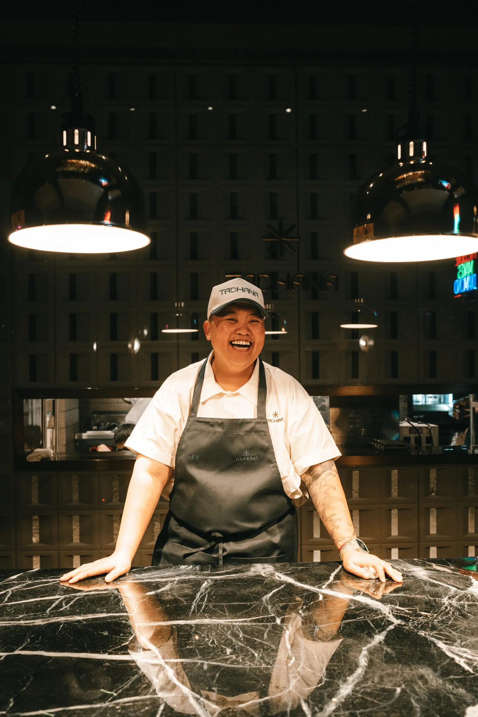 Smiling chef wearing a white shirt and black apron behind a dark marble counter in a kitchen, with warm overhead lights above.