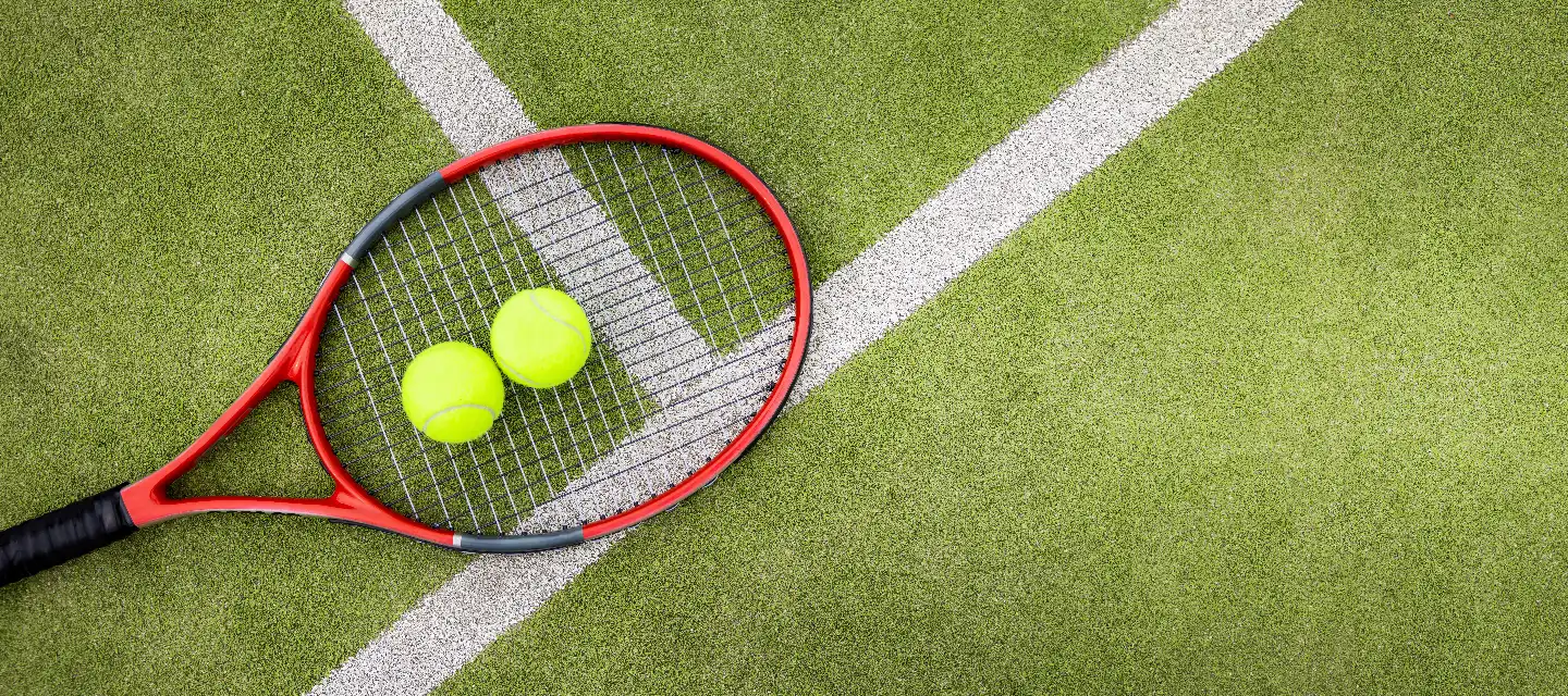 Red tennis racket with two neon-yellow tennis balls resting on the strings, on a green grass court near white boundary lines.