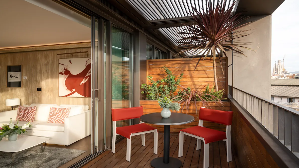 Balcony seating area with a round black table, two red chairs, and potted plants along a wooden screen; sliding glass door to the living room.