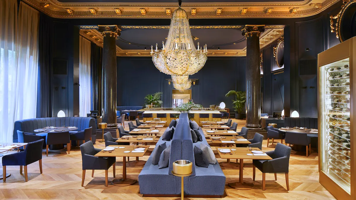 Luxurious dining room with blue upholstered seating, long wooden tables, and a grand chandelier centerpiece.