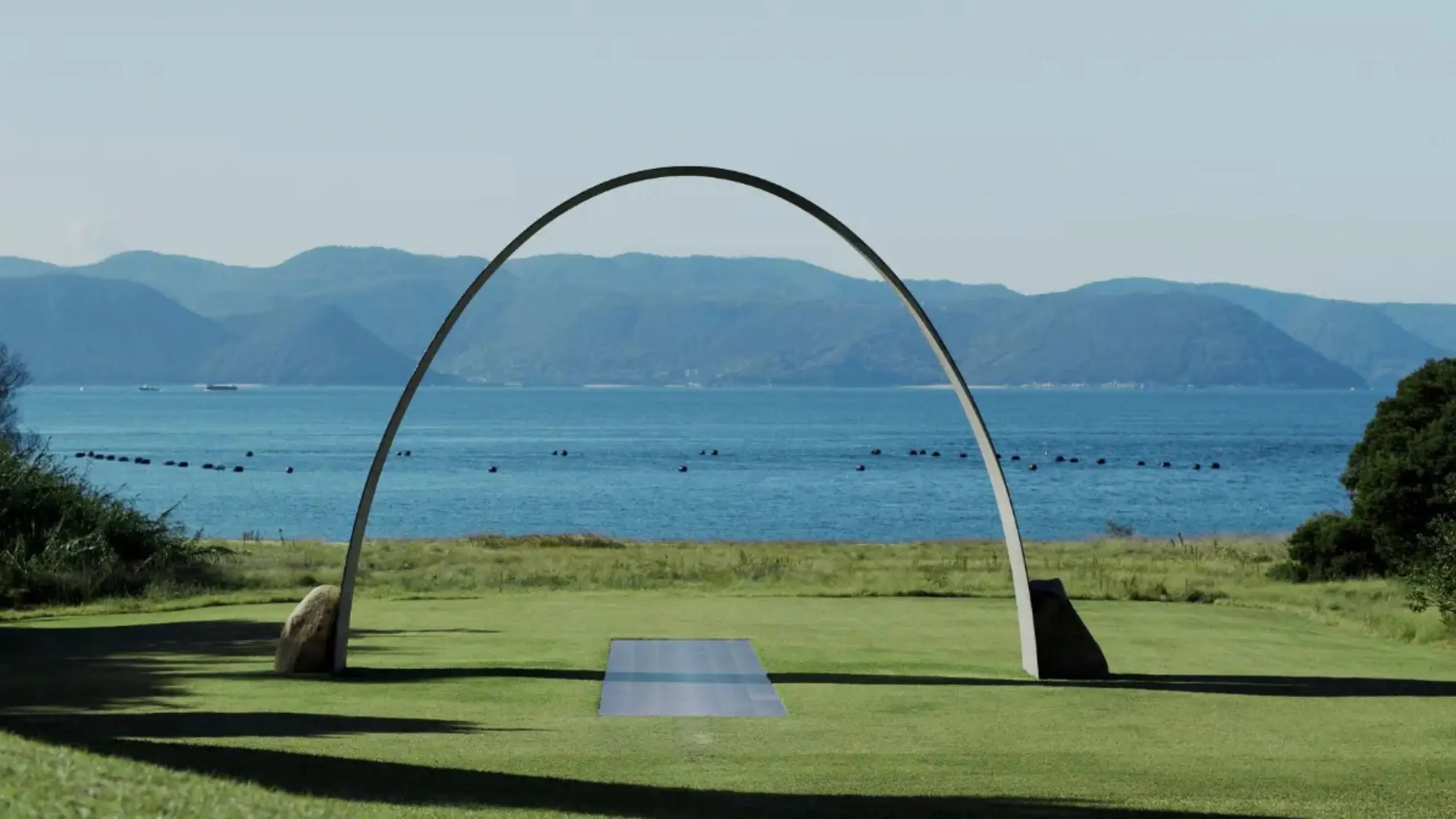 Curved metal arch sculpture over a grassy park facing a calm bay with distant mountains in the background.