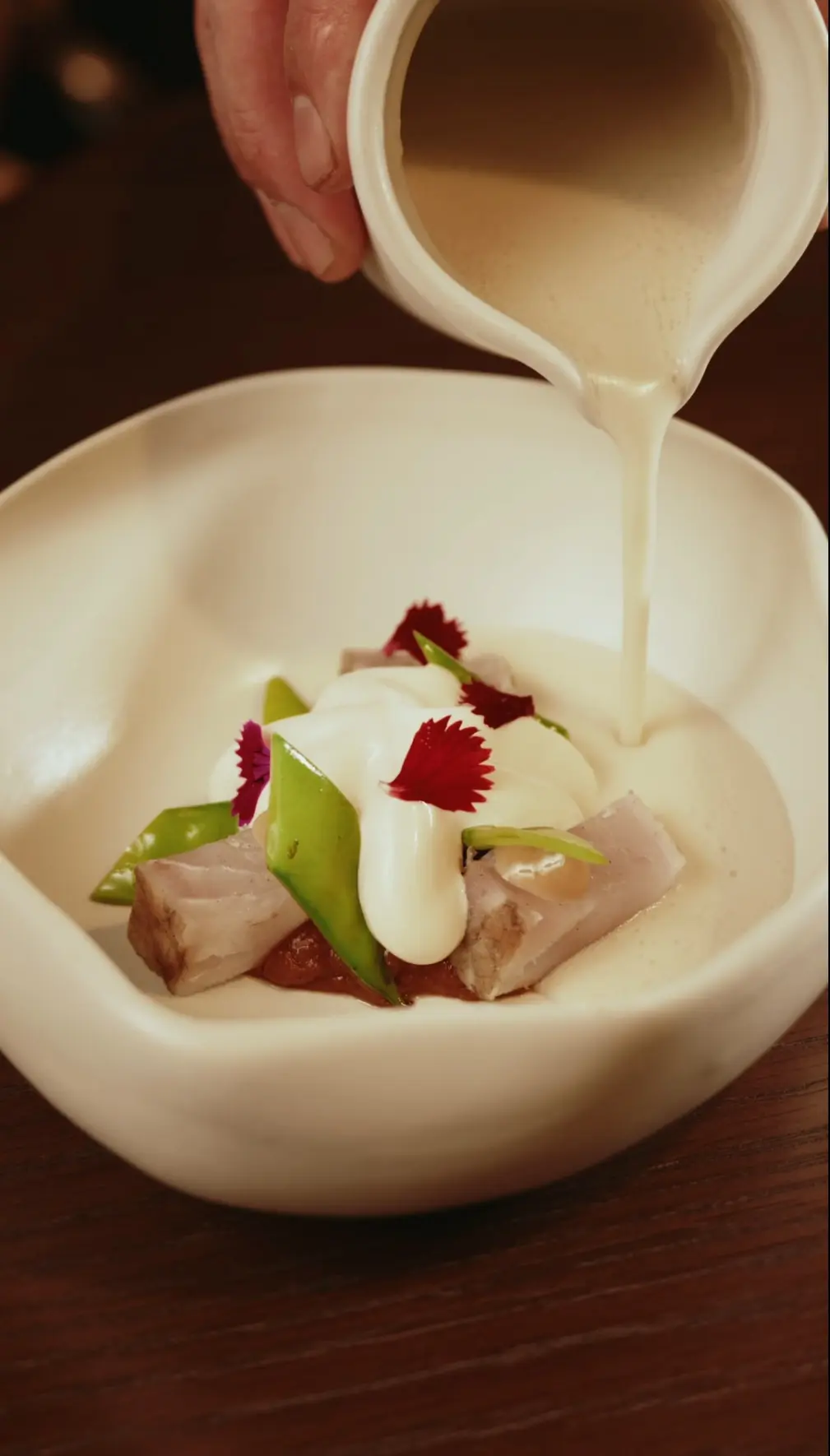 Creamy sauce is poured over a plated dish of diced pork, snap peas, greens, and red edible flowers in a white bowl.