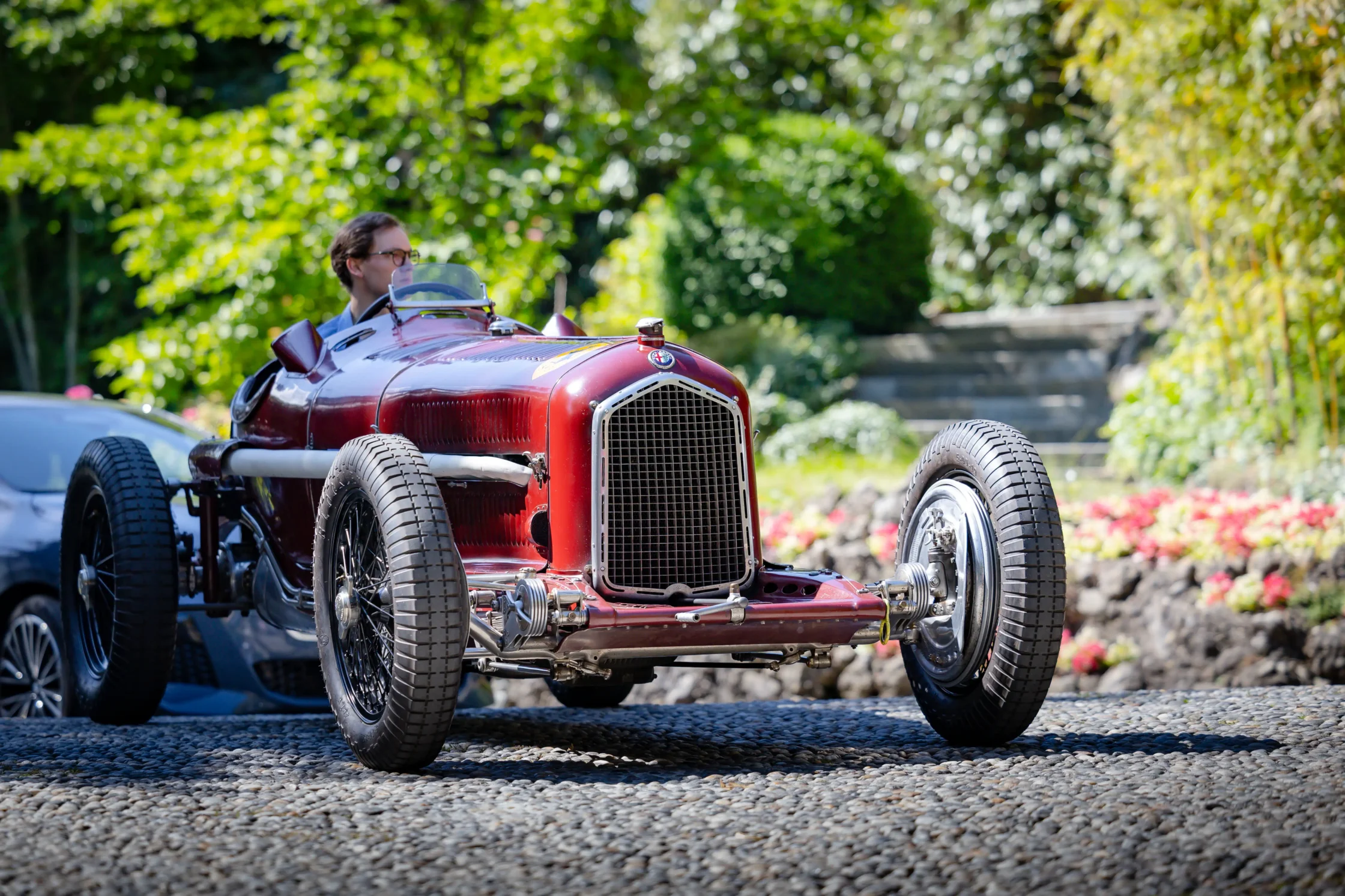 Red vintage race car on a trailer with its large front grille visible, driver in goggles in the cockpit, lush green background.