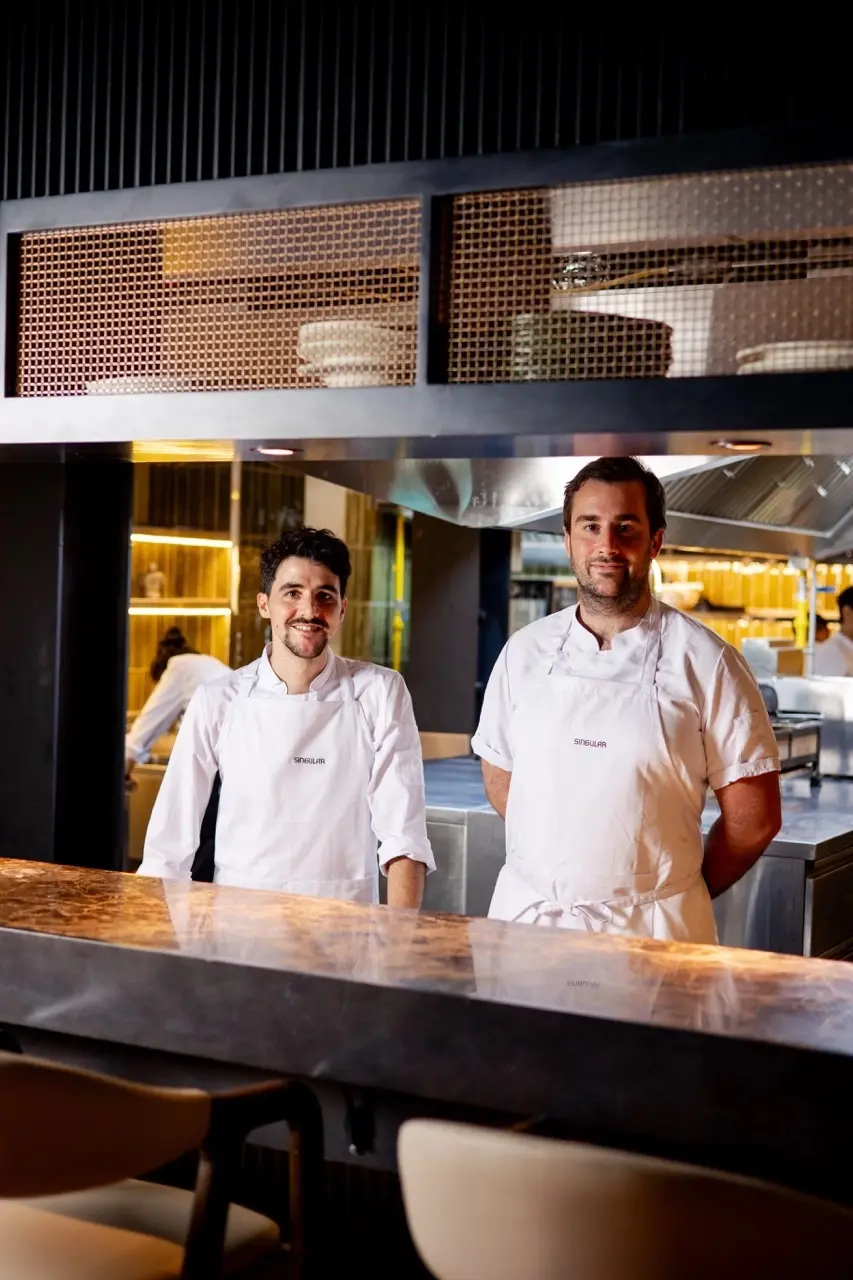 Two chefs in white aprons stand behind a marble kitchen counter in a modern restaurant kitchen, smiling at the camera.