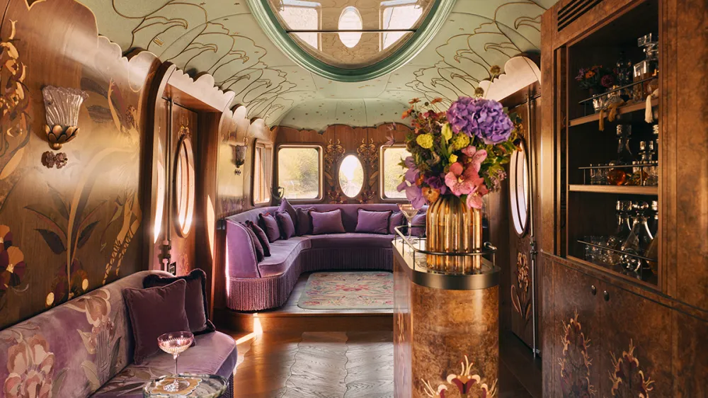 Lavish vintage train carriage with purple upholstered seating, ornate floral wallpaper, and a central flower arrangement on a marble-topped bar.