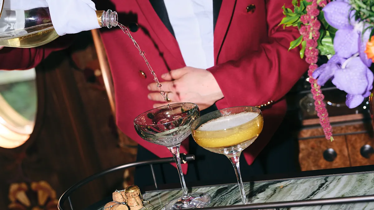 A bartender in a burgundy blazer pours champagne into two coupe glasses on a marble bar, with purple flowers nearby.
