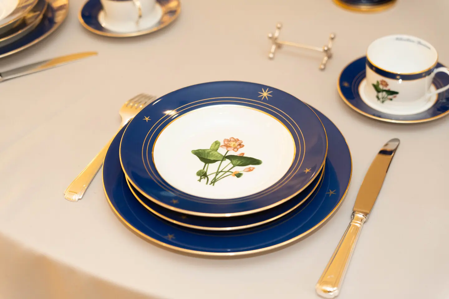 Elegant blue and gold table setting with nested plates and a floral center plate on a beige tablecloth.