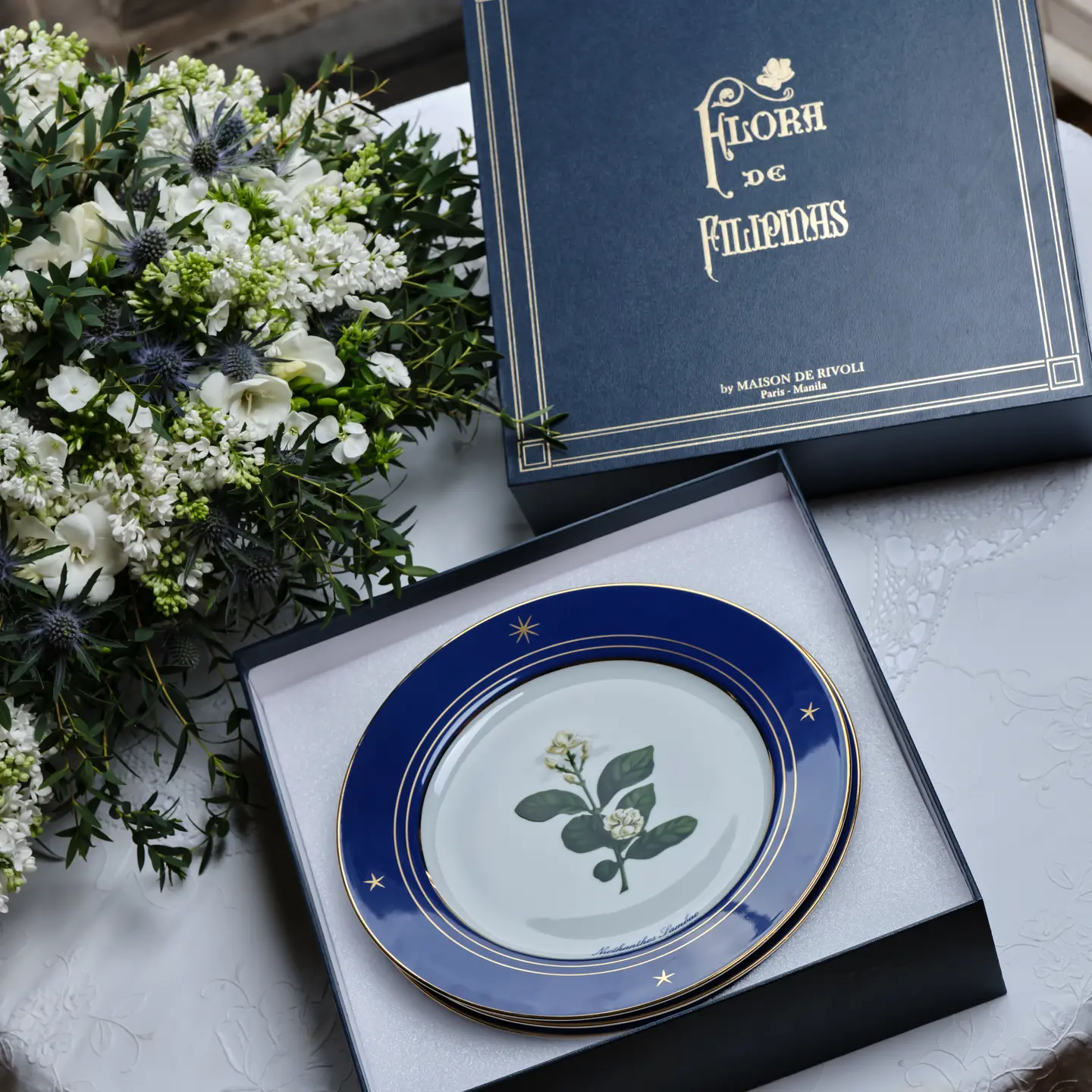 Blue gift box with gold lettering beside a navy-rimmed plate with botanical print, placed near a white bouquet of flowers as a present.