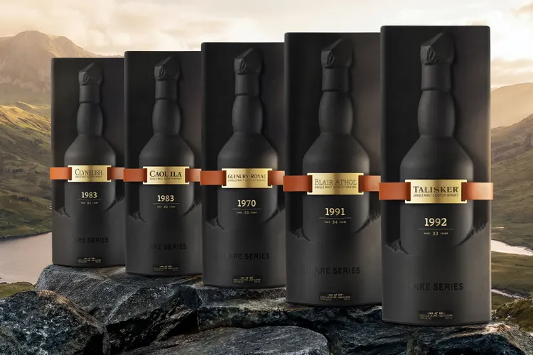Six dark bottles with copper bands lined up on a rocky ledge, a scenic mountain landscape in the background, part of a whisky collection.