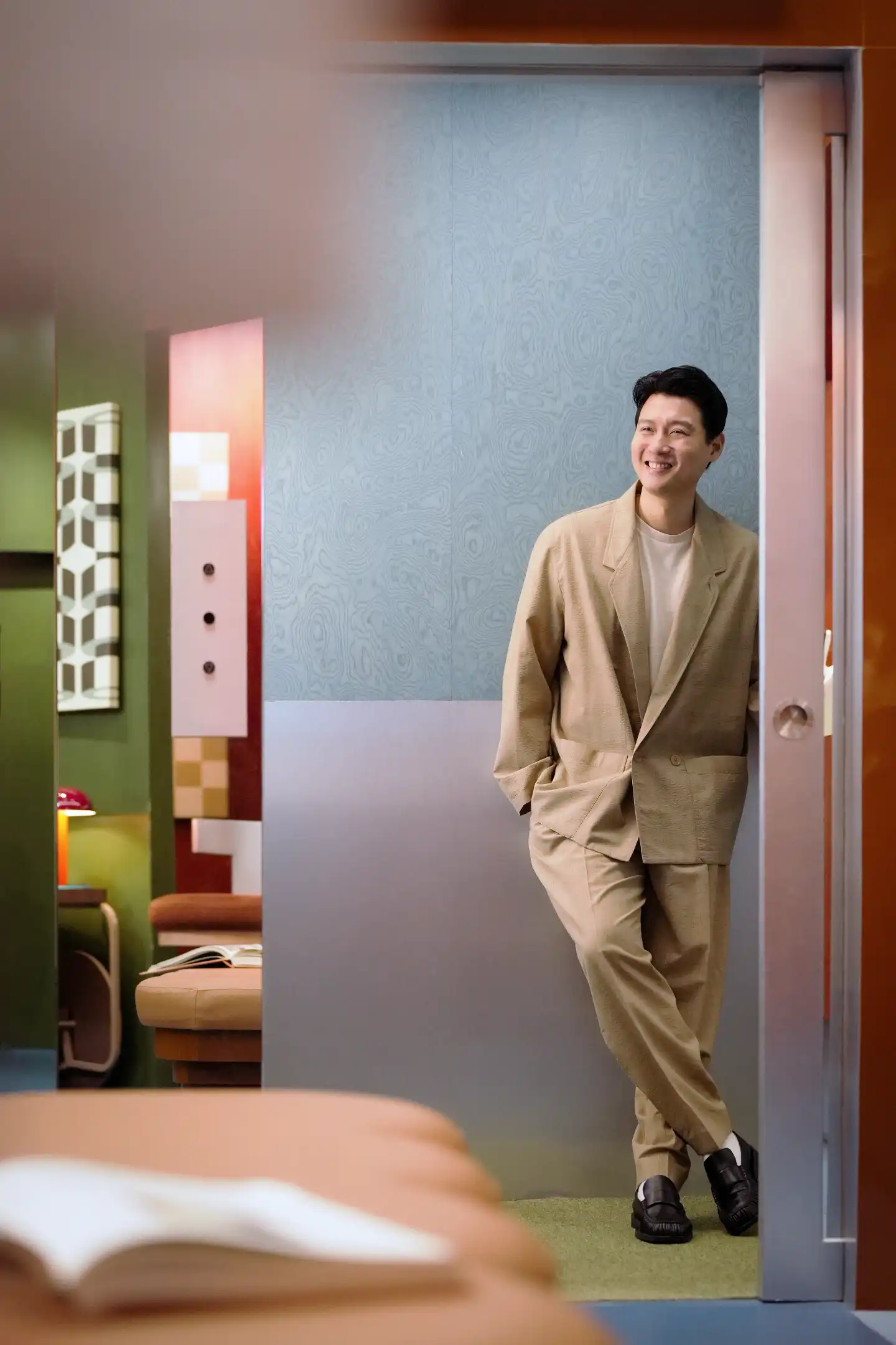 Man in a beige suit stands in a doorway, smiling in a retro-colored showroom with patterned walls and furniture visible in the background.